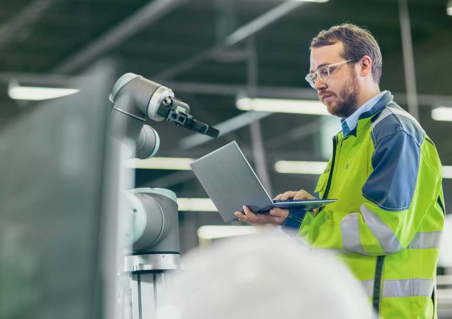 Ein Man mit gelb-grüner Sicherheitsjacke arbeitet mit einem Laptop in der Hand an einem Roboter