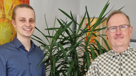 Tim, der bei AKQUINET sein duales Studium absolviert hat, steht neben Holger im Büro. Im Hintergrund ein Bild vom Atelier Lichtzeichen und eine Pflanze.